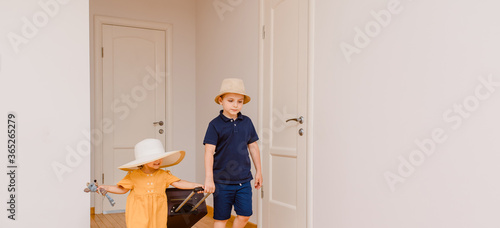 Happy girl and boy, brother and sister are carrying a big suitcase in room with white walls and wood floor