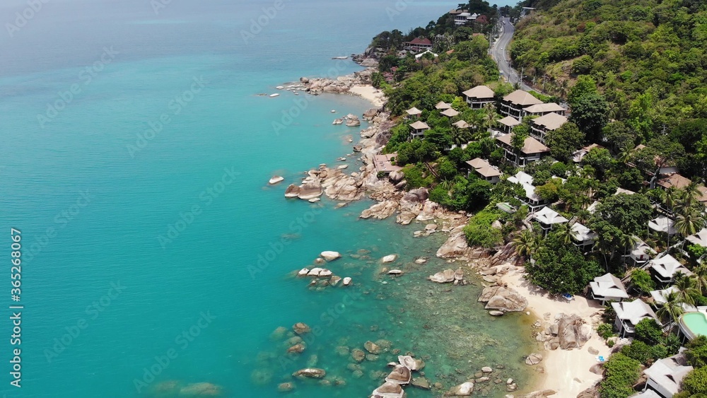 Small houses on tropical island. Tiny cozy bungalows located on shore of Koh Samui Island near calm sea on sunny day in Thailand. Volcanic rocks and cliffs drone top view.
