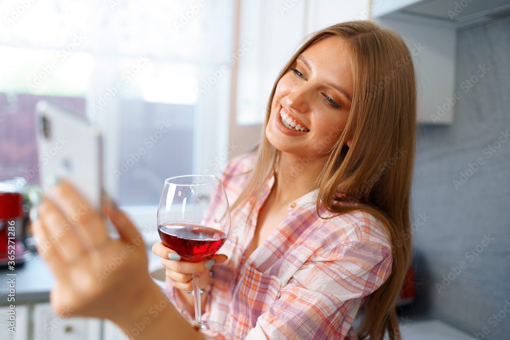 Happy relaxed young woman standing in kitchen with glass of red wine and using her smartphone Happy relaxed young woman standing in kitchen with glass of red wine and using her smartphone