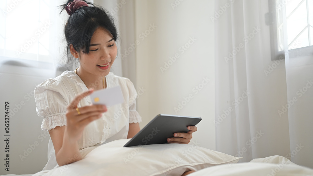 Asian woman is shopping online with a computer tablet while sitting on the bed in a comfortable bedroom. Asian woman is shopping online with a computer tablet while sitting on the bed in a comfortable bedroom.