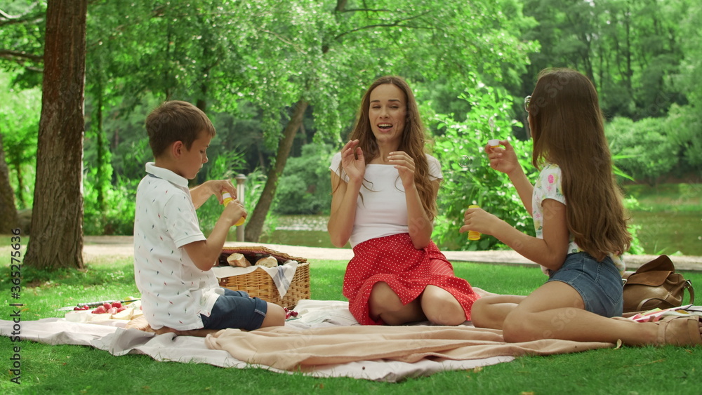 Girl and boy blowing bubbles in summer park. Smiling woman relaxing with kids Girl and boy blowing bubbles in summer park. Smiling woman relaxing with kids