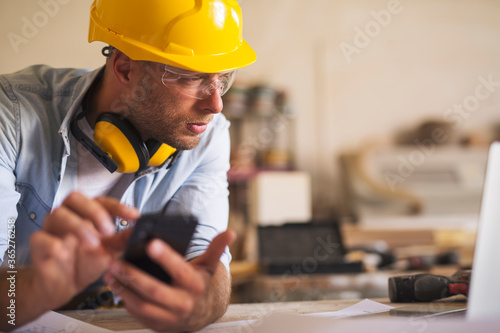 Close up of young carpenter using mobile phone while looking at laptop photo