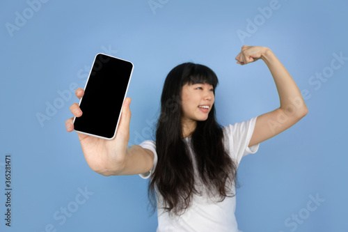 Showing phone with blank screen. Portrait of young asian woman isolated on blue studio background. Beautiful cute girl in casual. Human emotions, facial expression, sales, ad, online shopping concept.