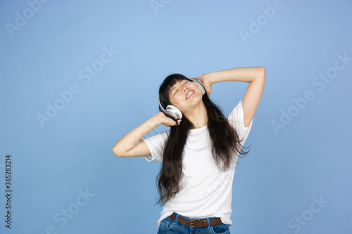 Listening to music in headphones. Portrait of young asian woman isolated on blue studio background. Beautiful cute girl in casual. Human emotions, facial expression, sales, ad, online shopping concept