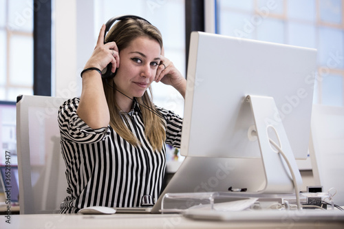 Giovane impiegata  bionda in camicia a righe seduta nella sua postazione lavorativa del suo ufficio di fronte lavora al computer indossando delle cuffie photo