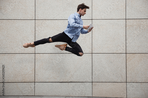 Jumping young buinessman in front of city building wall, on the run in jump high. Scrolling tablet, moving to daily routine inspired and sportive. Young ballet dancer in casual clothes and sunshine.