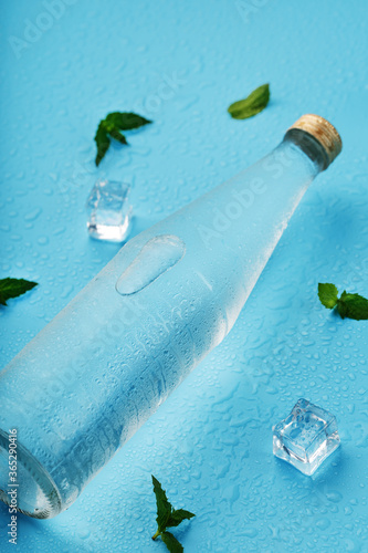 A bottle of ice water, ice cubes, drops and mint leaves on a blue background.