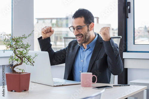 Manager uomo moro vestito con camicia blu e giacca nera porta degli occhiali da vista mentre lavora seduto nella postazione del suo ufficio ed esulta felice