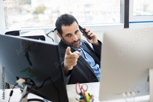 manager moro con giacca nera e camicia blu seduto nella sua postazione lavorativa tra due monito parla al telefono e gesticola con le mani photo