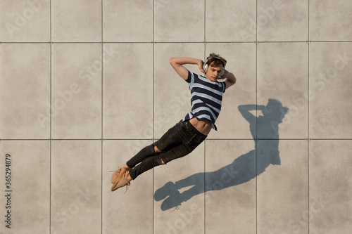 Jumping young buinessman in front of city building wall, on the run in jump high. Listening to music, moving to daily routine inspired and sportive. Young ballet dancer in casual clothes and sunshine.