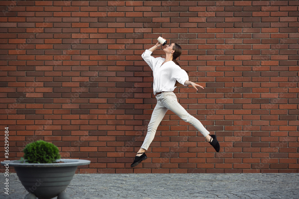 Coffee drinking. Jumping young woman in front of city building wall, on the run in jump high. Hurrying up to daily routine inspired and sportive. Young ballet dancer in casual clothes and sunshine.