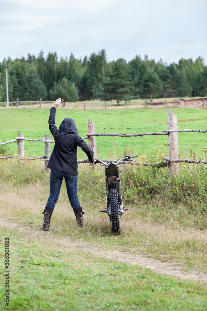 Rear view at female with rising hand standing in black raincoat with hood on head, dirt motorcycle on countryside road Rear view at female with rising hand standing in black raincoat with hood on head, dirt motorcycle on countryside road