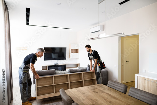 Moving with care. Full length shot of two contractor employees, young and aged worker in uniform moving personal belongings, furniture inside apartment. Moving service concept photo