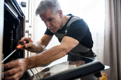 Close up shot of aged repairman in uniform working, fixing broken oven in the kitchen using screwdriver. Repair service concept
