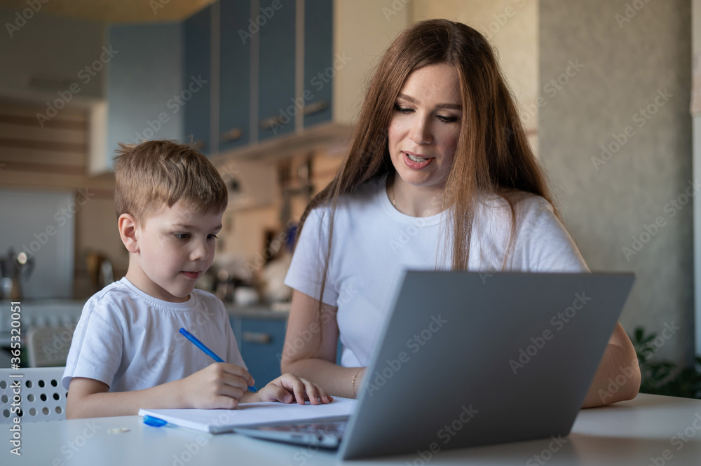 Caucasian woman helps boy learn lessons. The tutor checks the child's homework. Individual home-based quarantine. A diligent boy writes a dictation under the supervision of a teacher. Caucasian woman helps boy learn lessons. The tutor checks the child's homework. Individual home-based quarantine. A diligent boy writes a dictation under the supervision of a teacher.