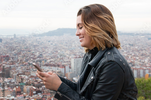 Fototapeta Naklejka Na Ścianę i Meble -  Girl smiles while looking at the mobile phone with the city in the background