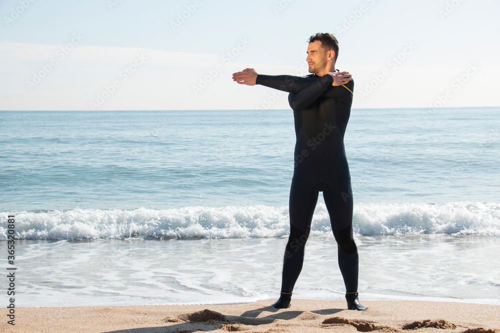 attractive and athlete guy in wetsuit stretches on the beach before entering the sea with the surfboard attractive and athlete guy in wetsuit stretches on the beach before entering the sea with the surfboard