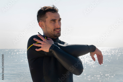 attractive and athlete  guy in wetsuit stretches on the beach before entering the sea with the surfboard