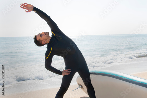 attractive and athlete  guy in wetsuit stretches on the beach before entering the sea with the surfboard