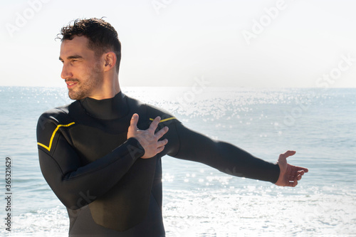 attractive and athlete  guy in wetsuit stretches on the beach before entering the sea with the surfboard