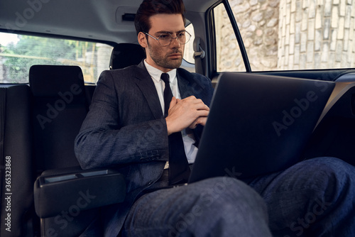 Confident young businessman seriously and thoughtfully looks at the laptop while sitting in the car. © Roman
