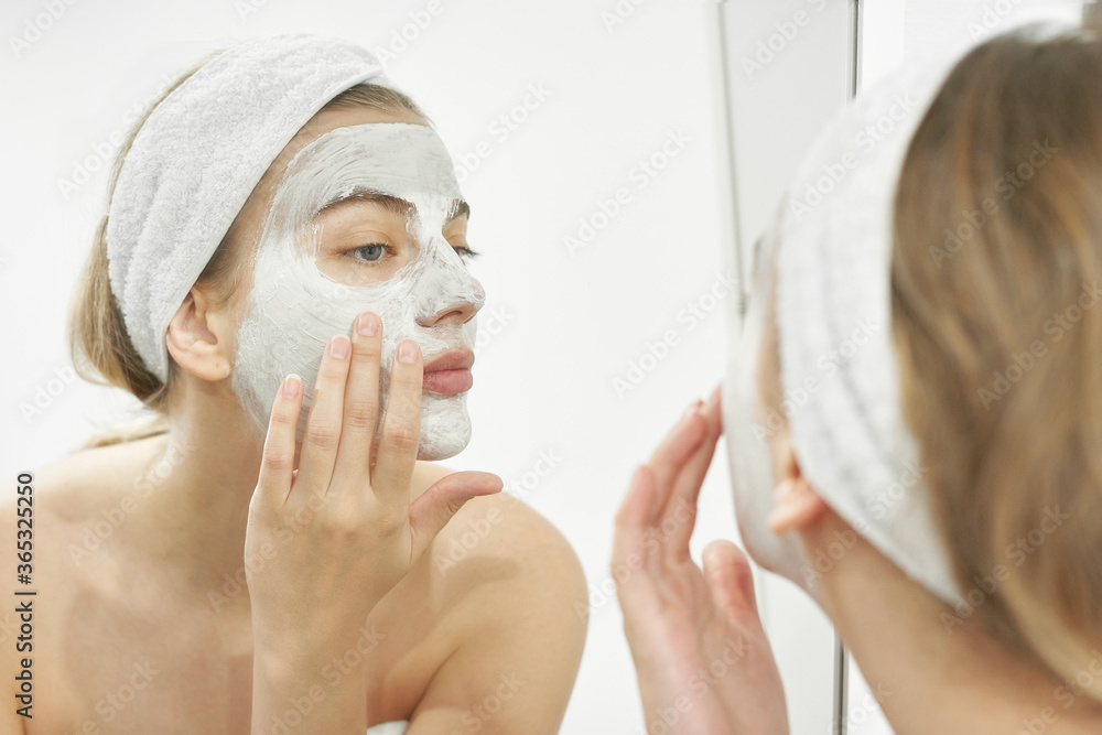 Beautiful European girl looks in the mirror and applies a white mask to her face. Summer home skin care