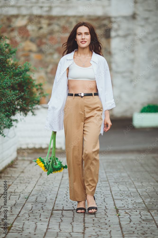 Young happy beautiful girl is walking in a shirt and pants with a bouquet of flowers of sunflowers on a city street