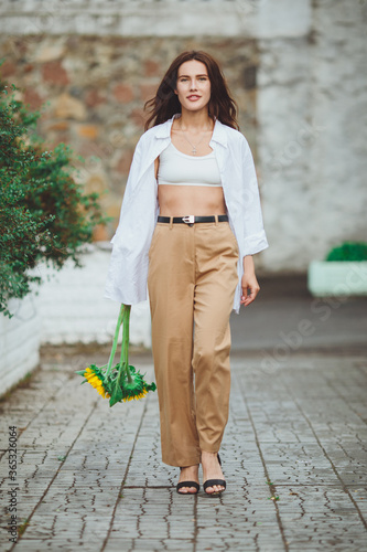Obraz na płótnie Young happy beautiful girl is walking in a shirt and pants with a bouquet of flowers of sunflowers on a city street w salonie