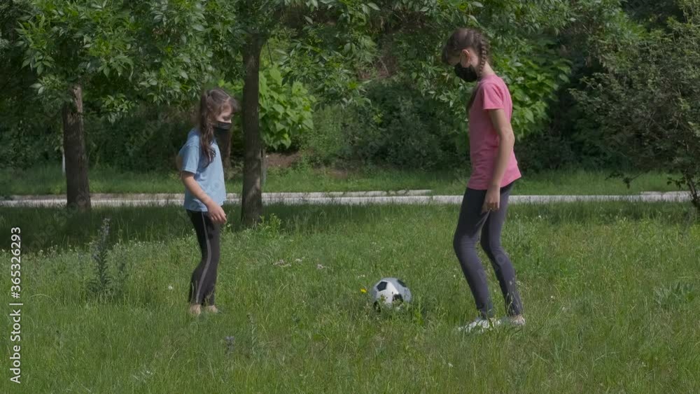 Sport outdoor in quarantine. Little girls in protective masks play with a soccer ball in a summer park on the lawn. Sport outdoor in quarantine. Little girls in protective masks play with a soccer ball in a summer park on the lawn.