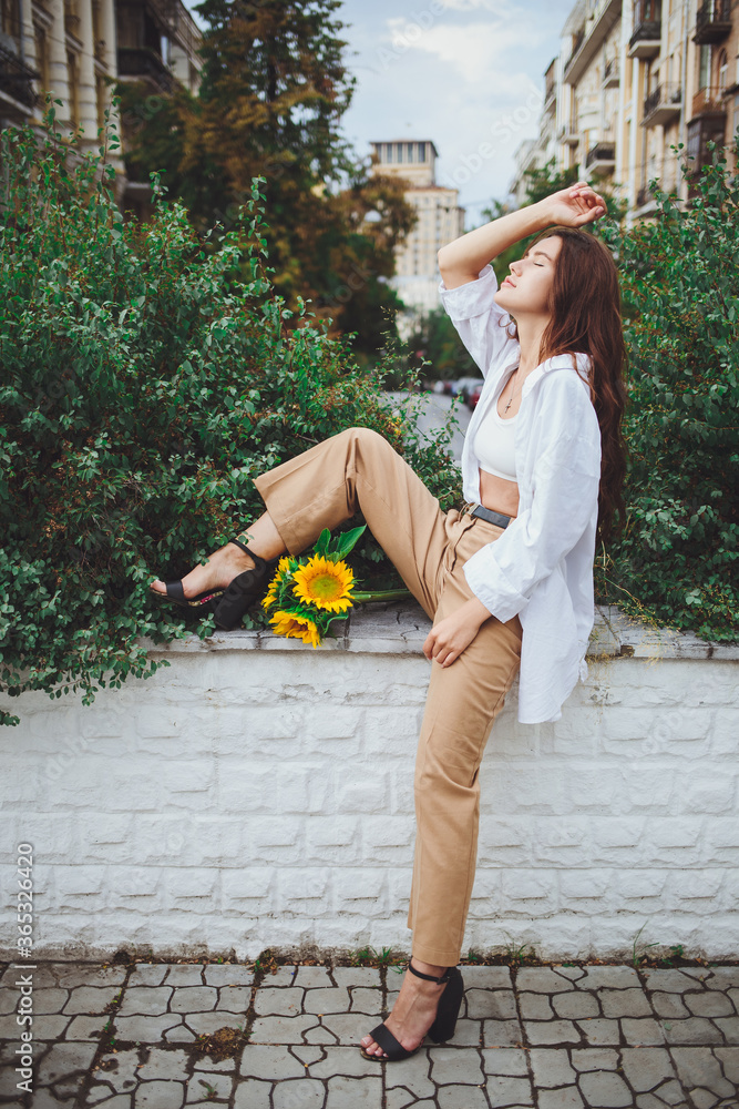 Young happy beautiful girl is walking in a shirt and pants with a bouquet of flowers of sunflowers on a city street