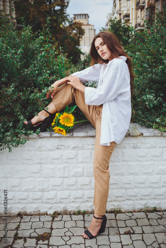 Young happy beautiful girl is walking in a shirt and pants with a bouquet of flowers of sunflowers on a city street