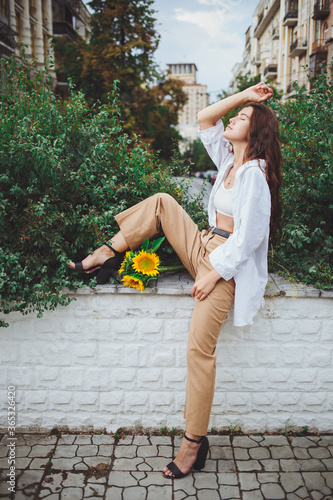 Obraz na płótnie Young happy beautiful girl is walking in a shirt and pants with a bouquet of flowers of sunflowers on a city street w salonie