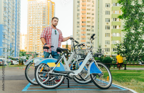 Fototapeta Naklejka Na Ścianę i Meble -  Street portrait of a bearded man in casual clothes standing on a bicycle parking lot and taking a bike for a walk, looking at the camera with a serious face. Adult man uses bicycle rental.