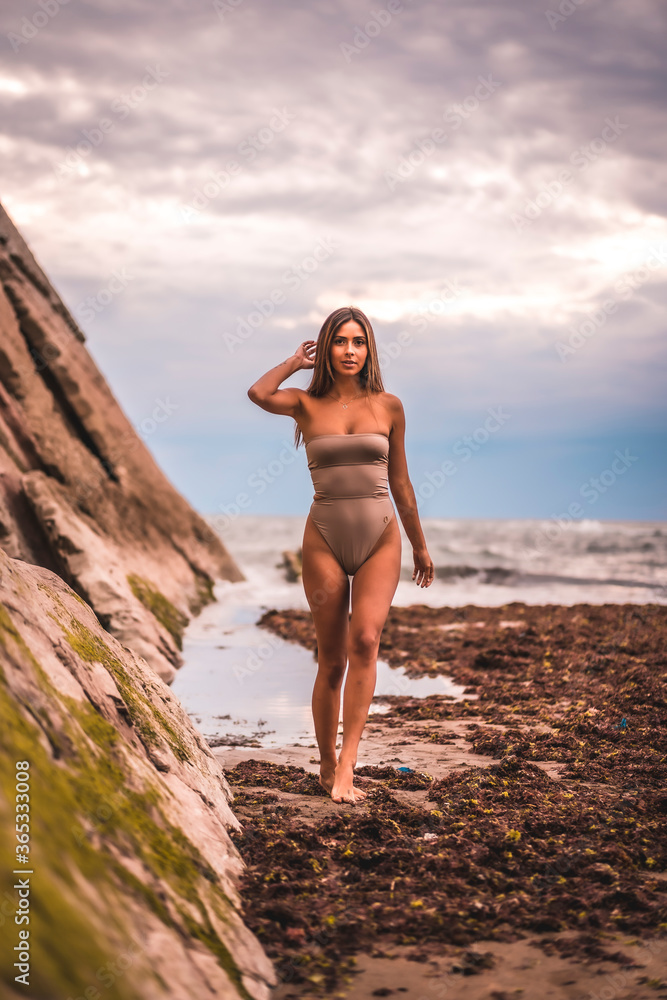 A dark-haired caucasian woman in a brown swimsuit on a natural background by the sea in the town of Zumaia, Gipuzkoa. Basque Country. In the summer sunset. Walking on some rocks on the in coast