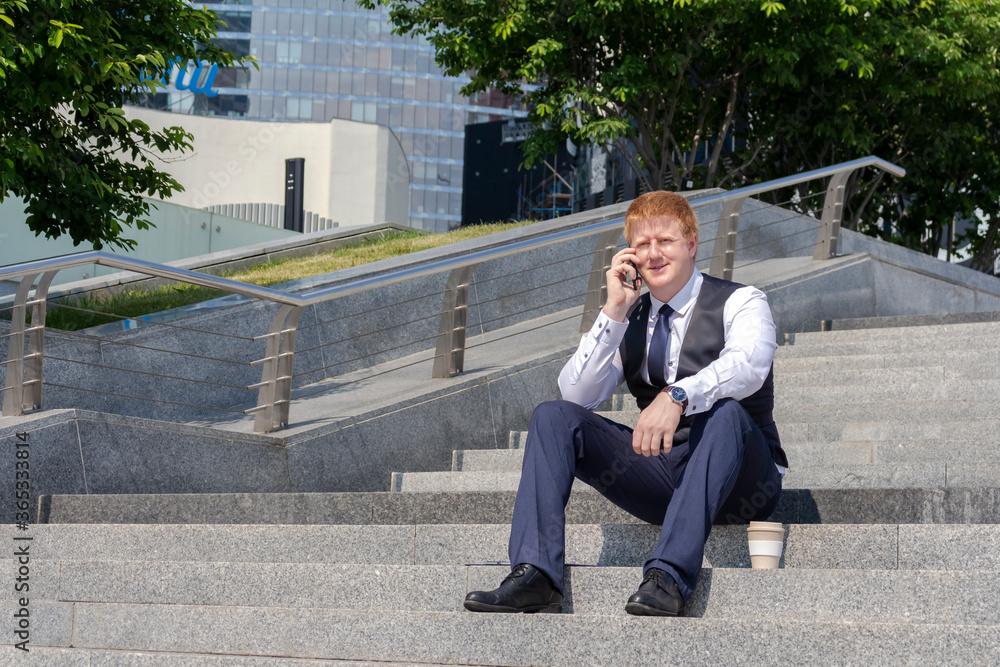 Adult caucasian man sits on the steps and talks on the phone. There is a reusable coffee cup next to him. The man is wearing a business uniform. Coffee break theme. Adult caucasian man sits on the steps and talks on the phone. There is a reusable coffee cup next to him. The man is wearing a business uniform. Coffee break theme.