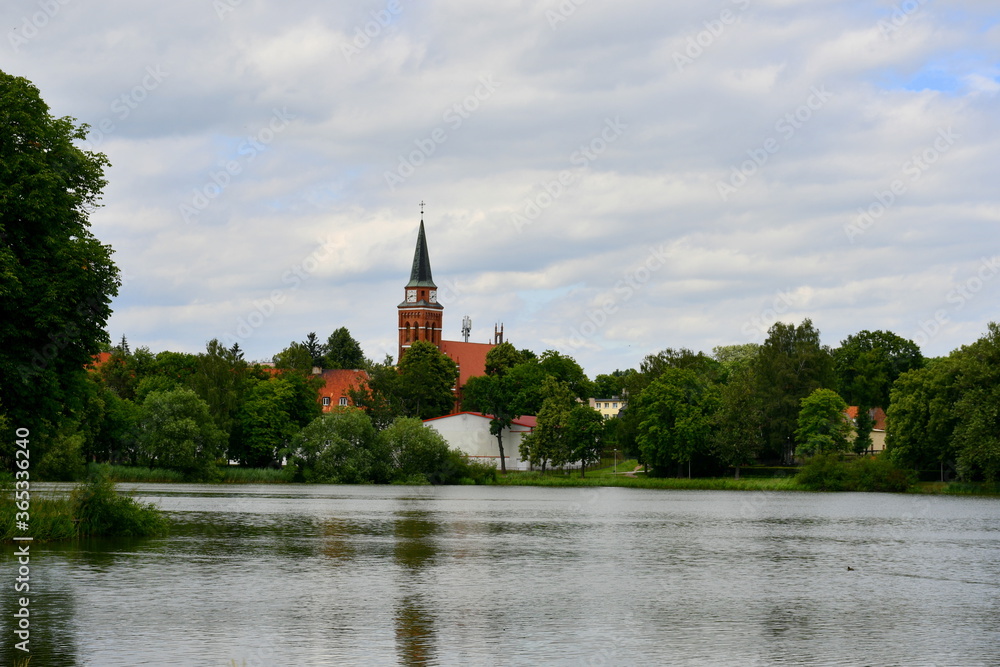 View of a shallow yet vast lake or river surrounded from all sides with dense trees and a big red brick church with a clock tower visible on the other side of the reservoir spotted in Poland