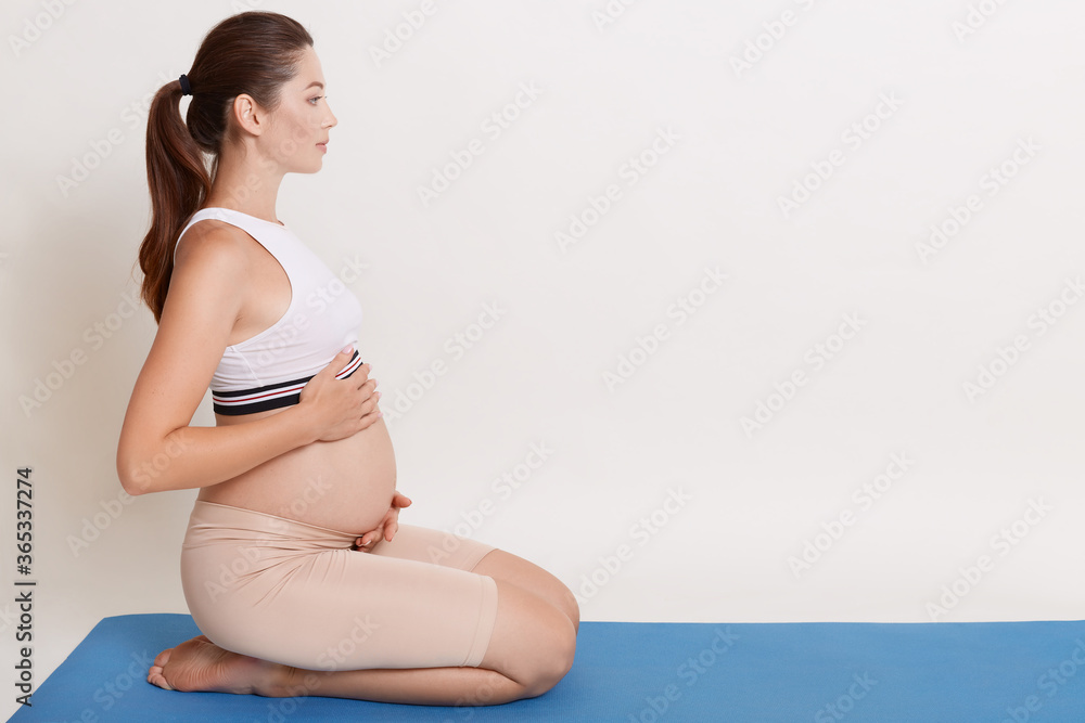 Beautiful expectant mother doing yoga at home, touching her bare belly, looking straight ahead, posing against white background, expectant mother relaxing after pregnancy gymnastic. Beautiful expectant mother doing yoga at home, touching her bare belly, looking straight ahead, posing against white background, expectant mother relaxing after pregnancy gymnastic.