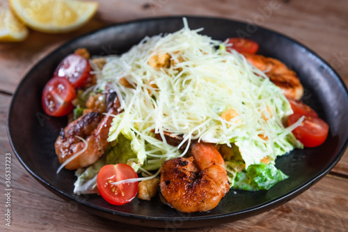 Appetizing Caesar salad with shrimps in a black plate on a wooden table next to a lemon. The concept of healthy food and serving