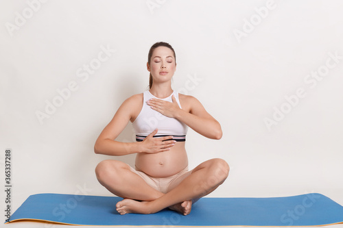 Pregnant woman doing yoga sitting in lotus position indoors. Future mother practice breathing exercises with one hand on pregnant belly and chest, keeping eyes closed.