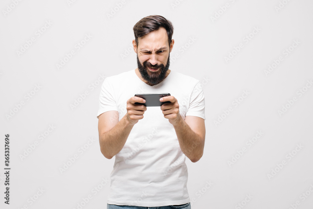 Expressive guy in a white T-shirt plays video games on a smartphone, looks at the screen and reacts emotionally. Young man plays a game on the phone, the focus is on a smartphone in his hands Expressive guy in a white T-shirt plays video games on a smartphone, looks at the screen and reacts emotionally. Young man plays a game on the phone, the focus is on a smartphone in his hands