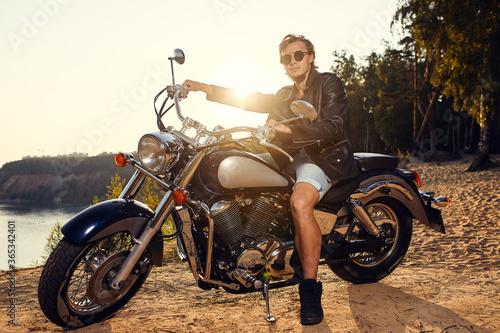 Brutal young man in sunglasses, blue jeans and a black leather jacket sitting on the custom motorcycle © Georgii