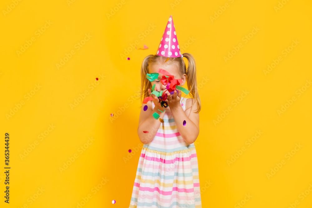 Happy birthday child girl in pink cup blowing confetti on colored yellow background. Celebration, childhood. Happy birthday child girl in pink cup blowing confetti on colored yellow background. Celebration, childhood.