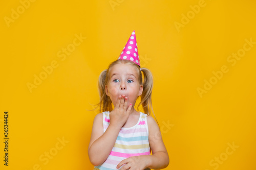 Happy birthday child girl in pink cap on colored yellow background. Toddler her mouth with hand.