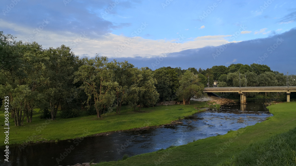View of shallow river with its both coasts covered with grass and a small concrete bridge connecting two sides of a road leading through a forest View of shallow river with its both coasts covered with grass and a small concrete bridge connecting two sides of a road leading through a forest
