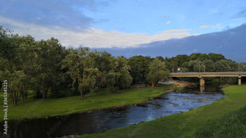 View of shallow river with its both coasts covered with grass and a small concrete bridge connecting two sides of a road leading through a forest