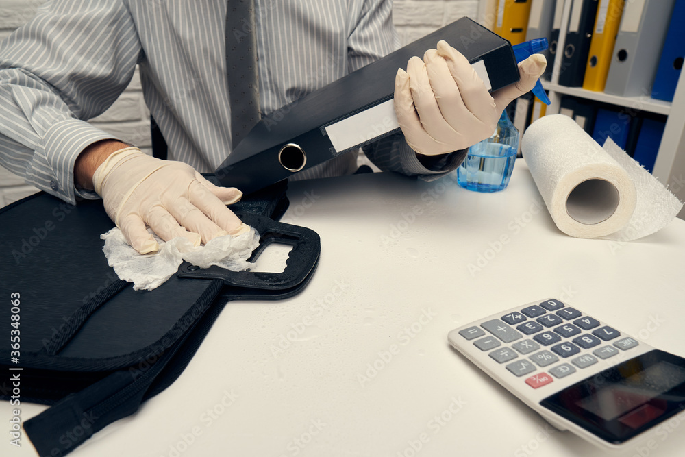 concept of cleaning or disinfecting the office desk - a businessman cleans the workplace, computer keyboard, document folders, uses a spray gun sanitizer, gloves and paper napkins.