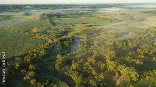 Aerial view of rural landscape with lush greenery, trees, river in the foreground and farmhouse, farmland in the background. American midwestern countryside scenery in the early sunny morning, summer