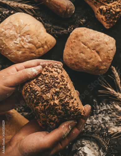 Hands holding loaf of bread on dark rustic background with sackcloth, flour, spikelets, rolling pin. Pastries and bread in a bakery, top view