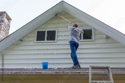 Man stands on porch roof to wash house with brush on extension