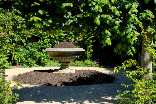 A very large weathered white stone planter urn topped up with soil in preparation for planting with flowers, at the base a circular patch of ground has been freshly dug in readiness for planting.